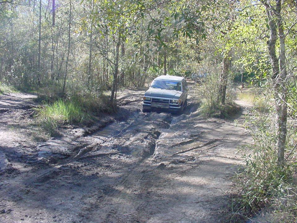 Silver compact pickup with camper shell driving through a deep mud hole on a wooded trail