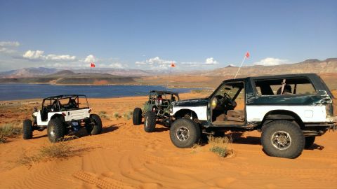 1994 Ford Bronco - Modified off-road vehicles are parked on sand dunes near a lake.