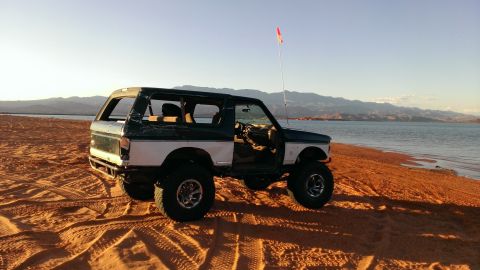 1994 Ford Bronco - A modified Ford Bronco sits on a sandy shore next to a body of water with mountains in the background.