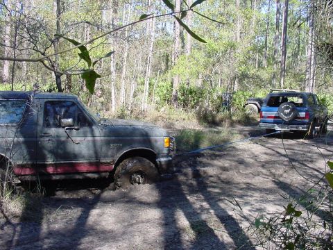 1991 Ford Bronco - A muddy 1991 Ford Bronco is being towed out of the mire by another SUV in a wooded area.