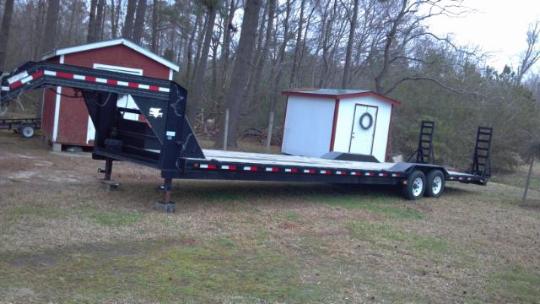 Gooseneck flatbed trailer parked in a grassy area with trees and two small buildings in the background