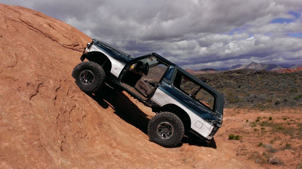 Ford Bronco climbing a steep red rock hill, doors open, with a cloudy sky