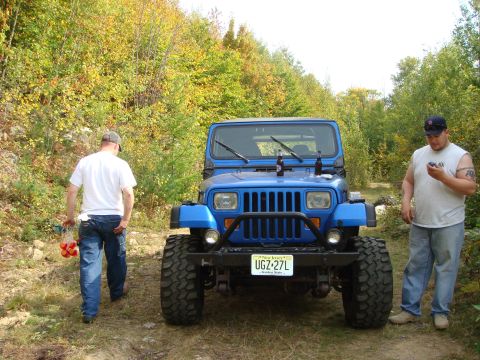 1990 Ford Bronco - A blue Jeep Wrangler is parked on a dirt path surrounded by trees, with two men standing near it.