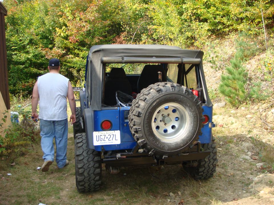 Blue Jeep Wrangler rear view with spare tire, license plate UGZ-27L, and a man walking away