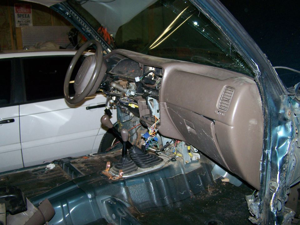 Interior of a disassembled vehicle with exposed wiring and dashboard components. Another vehicle is visible in the background