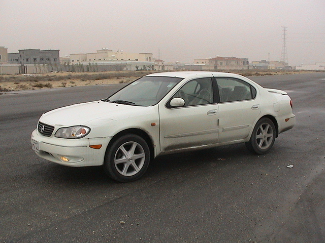 White four-door sedan with alloy wheels and dented lower body on empty desert asphalt lot