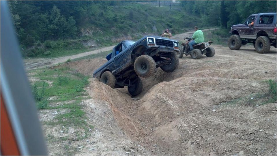 Lifted dark Ford Bronco climbing steeply out of a dirt gully at an off-road park, ATV and truck nearby