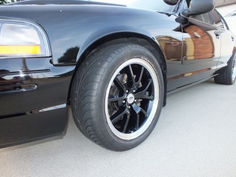 2003 Ford Crown Victoria - Close-up of a black sedan's front wheel and fender, showing aftermarket rims and tires.
