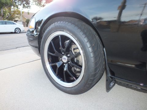 2003 Ford Crown Victoria - A black Ford Crown Victoria's front wheel and fender are shown with aftermarket rims and tires, with another white car in the background.