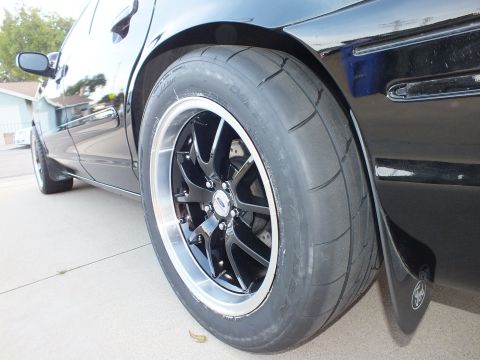 2003 Ford Crown Victoria - Close-up of a black car's rear wheel a performance tire and aftermarket alloy wheel, with a Ford logo visible on the mud flap.