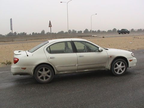 2004 Nissan Maxima - Side view of a white 2004 Nissan Maxima parked on a wet road, with a pickup truck visible in the background.