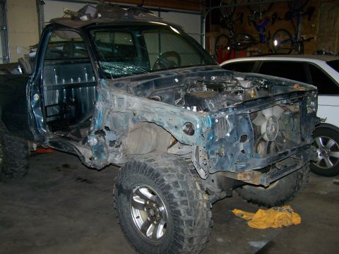 1988 Suzuki Samurai - A heavily damaged teal pickup truck sits in a garage, missing its front end and with a shattered windshield.