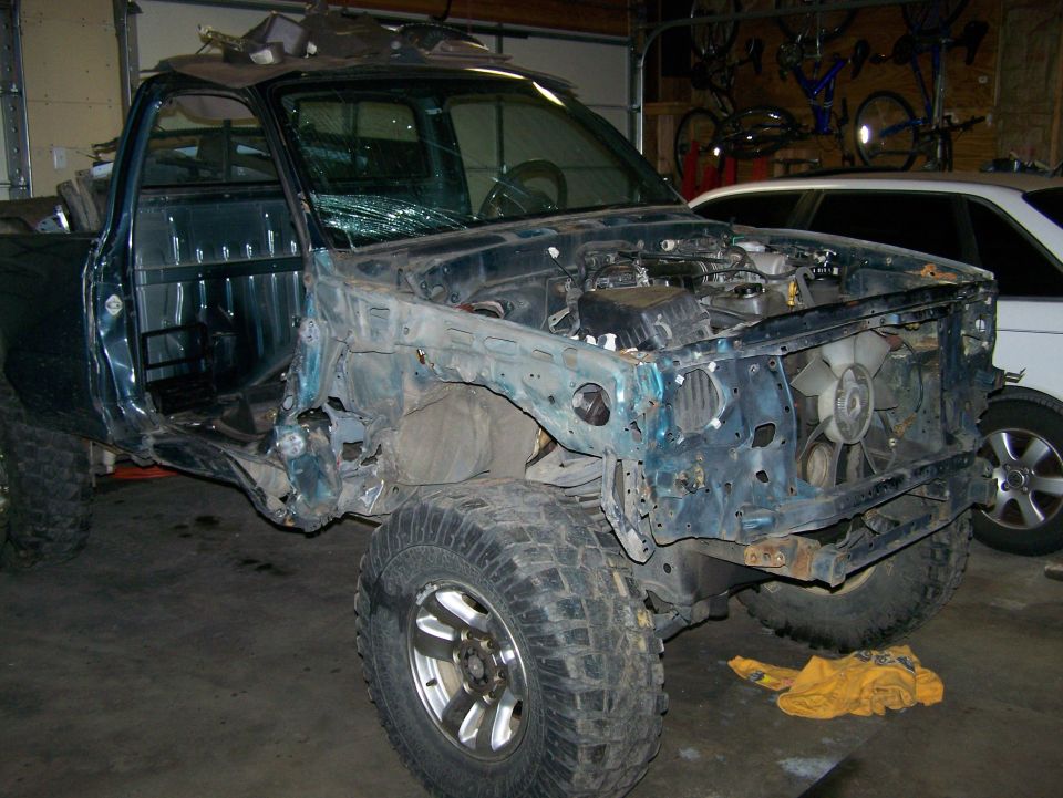 Damaged teal pickup truck with front end removed, open doors, and broken windshield