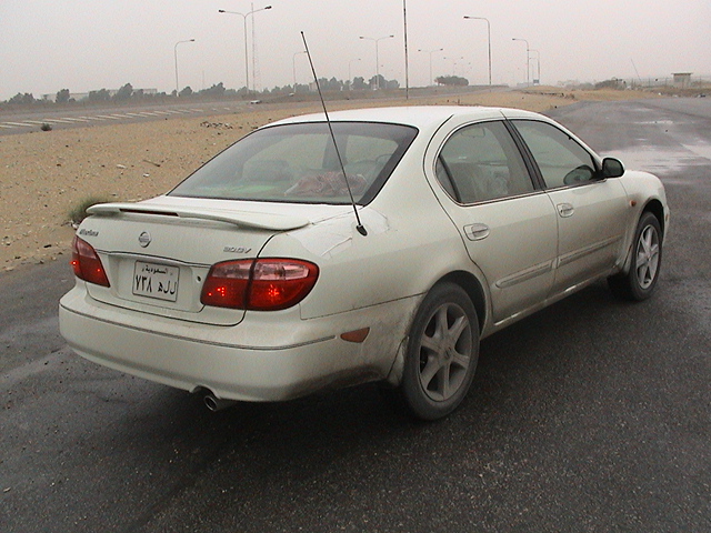 White Nissan sedan, rear 3/4 view, dirty, with a spoiler and a license plate