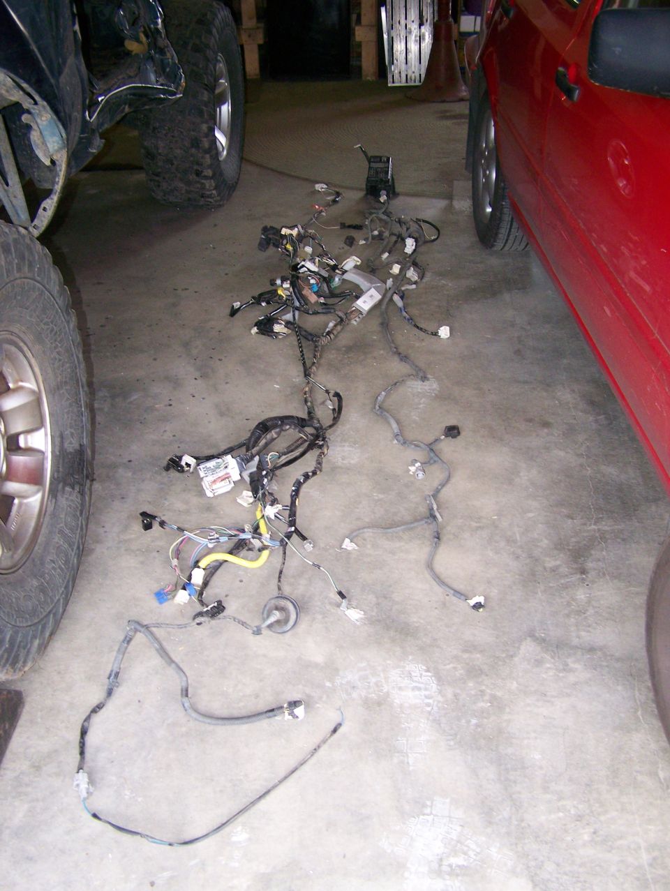 Vehicle wiring harness spread out on concrete floor next to a red vehicle and a dark-colored vehicle