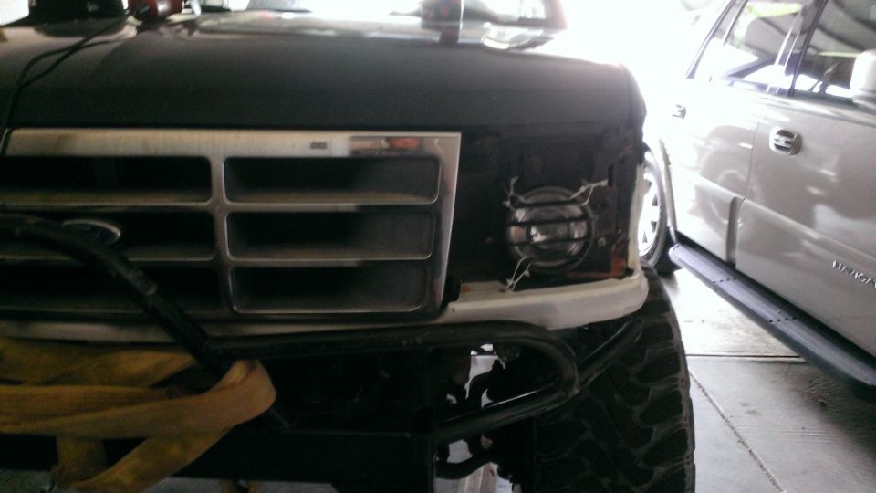 Front grille and headlight area of a dark-colored Ford Bronco, possibly undergoing repair. Another white SUV is parked to the right