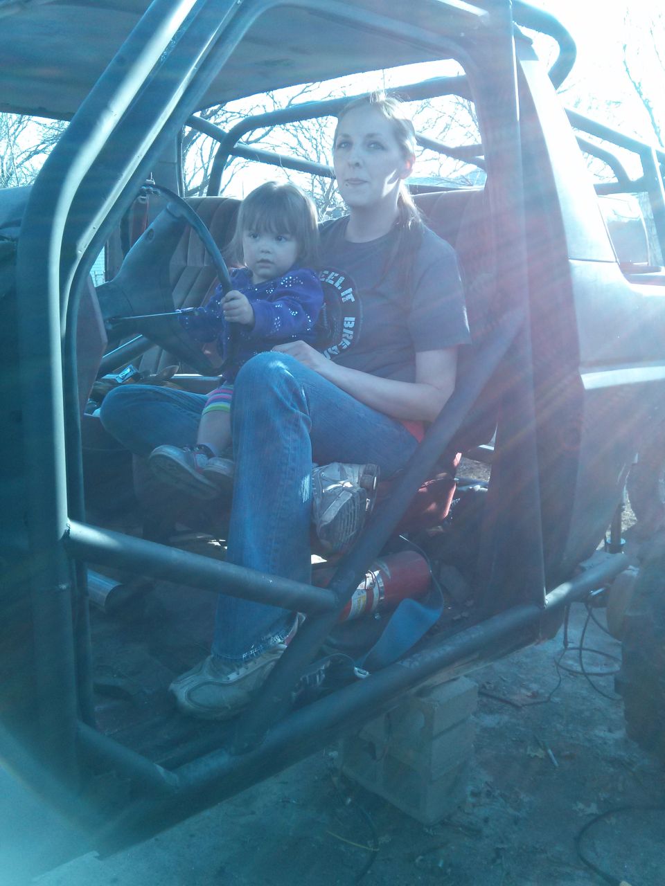 Woman holding a child inside a vehicle with a roll cage and exposed interior. The vehicle appears to be a modified SUV