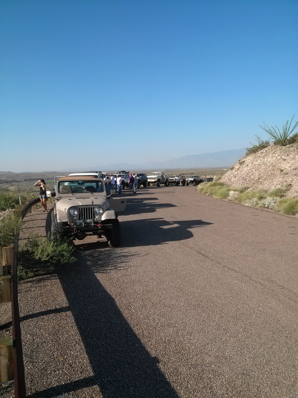 Tan Jeep CJ-7 parked on gravel roadside, door open. Line of other vehicles behind it. Desert landscape