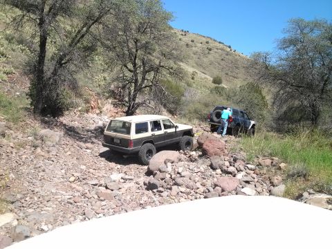 1981 Ford F-150 - Two SUVs are seen traversing a challenging rocky terrain on an off-road trail under a bright blue sky.