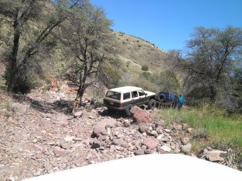 1981 Ford F-150 - Two off-road vehicles navigate a challenging, rocky trail in a dry, hilly landscape under a clear blue sky.