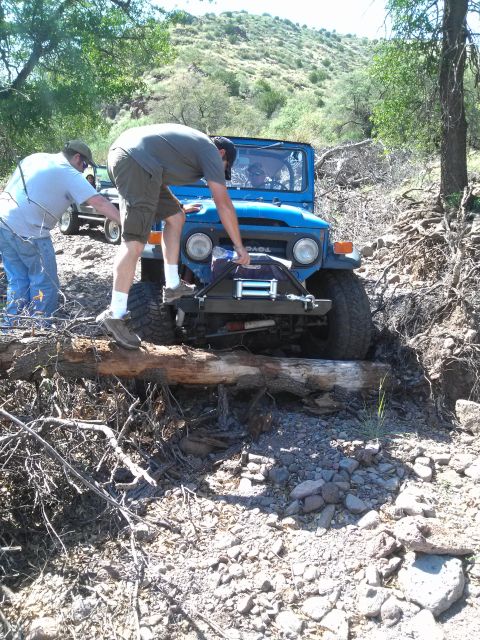 1981 Ford F-150 - Two people assist a blue Toyota Land Cruiser FJ40 as it drives over a log on a rough, wooded trail.