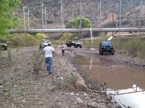 1981 Ford F-150 - Off-road vehicles gather near a creek and bridge, with water spraying from an overhead pipe.