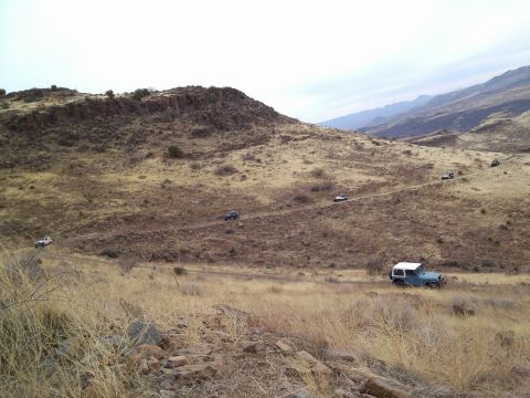 1981 Ford F-150 - A convoy of off-road vehicles navigates a winding dirt path through a , dry mountain terrain.