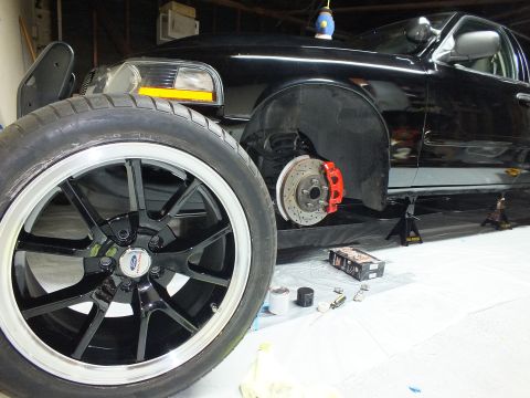 2003 Ford Crown Victoria - Garage shot of a black sedan on jack stands showing a red caliper, drilled rotor and a wheel set aside.