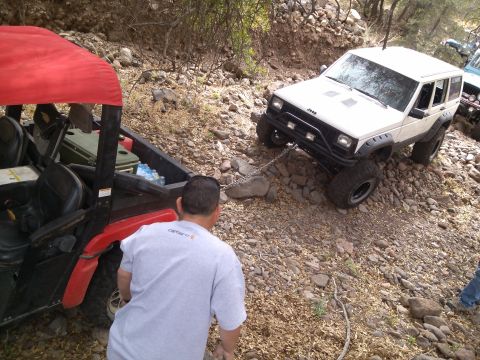 1981 Ford F-150 - A white Jeep Cherokee is being towed by a UTV on a rocky, off-road trail.