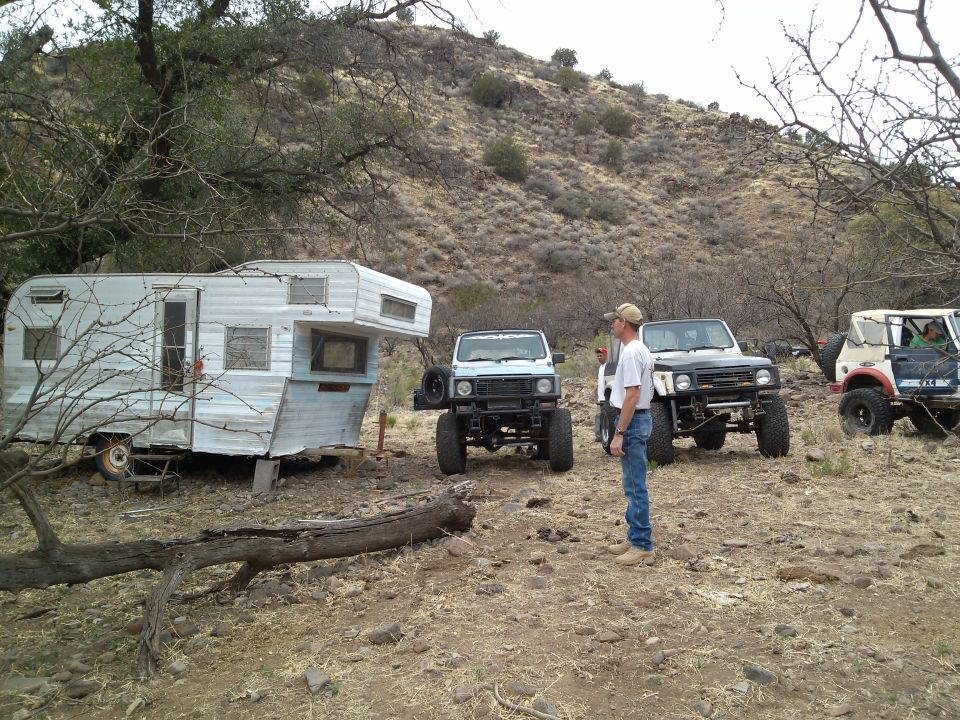 Three off-road vehicles and a trailer parked on a dirt lot with a hill in the background