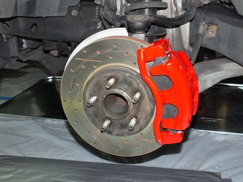 2003 Ford Crown Victoria - Underside view of a red front caliper clamping a drilled-slotted rotor on a five-lug hub.