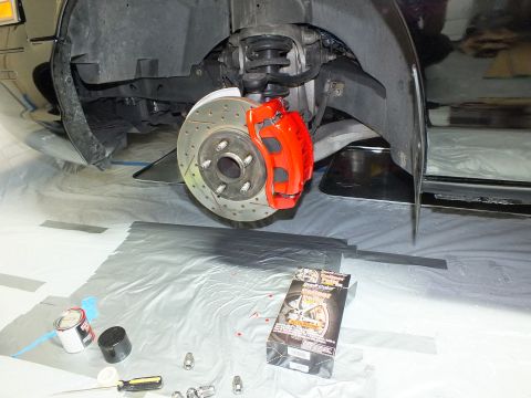 2003 Ford Crown Victoria - Front brake upgrade in progress: red caliper, drilled rotor, with paint can and parts box on plastic-covered floor.
