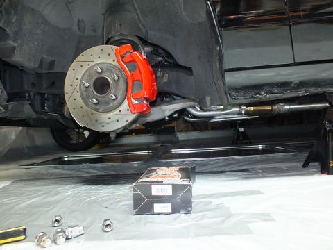 2003 Ford Crown Victoria - Underbody view of red caliper and drilled rotor beside exhaust piping and a black parts box on the floor.