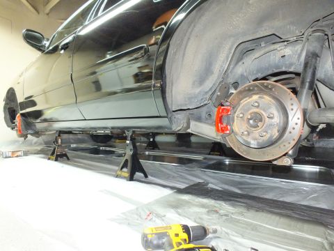 2003 Ford Crown Victoria - Black sedan elevated on jack stands above floor plastic, drilled rotor visible and a yellow drill nearby.