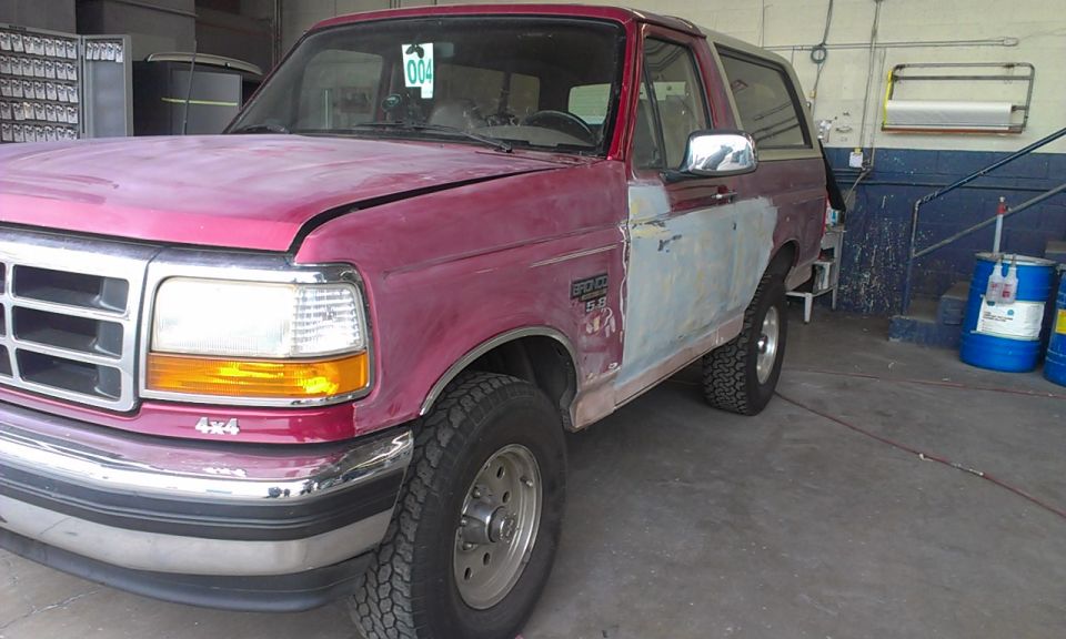 Red Ford Bronco with primer on the passenger side door, parked indoors