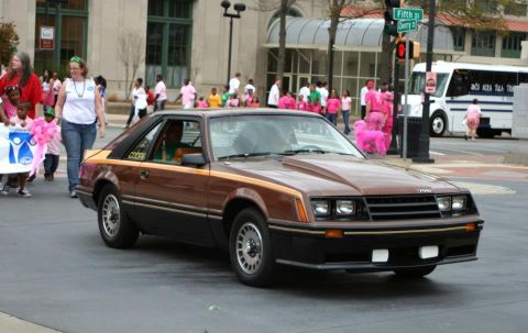1980 Ford Mustang - A brown 1980 Ford Mustang is seen driving in a parade with spectators on the sidewalk.