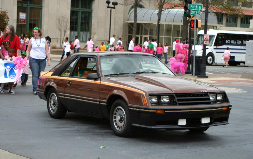 Brown 1980 Ford Mustang driving down street with people walking nearby