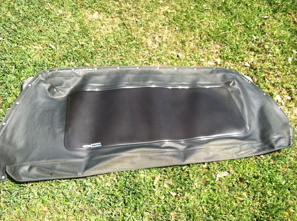 Black soft top with a clear plastic rear window, laid out on grass. Small logo visible on the window frame