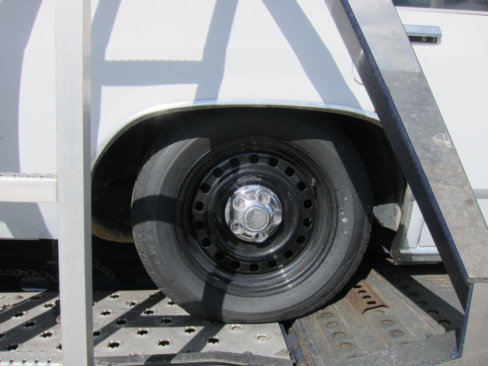 Close-up of a car's rear wheel and fender, likely on a trailer. The wheel has a black steel rim with a chrome hubcap
