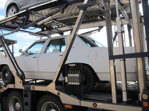 1991 Ford LTD Crown Victoria - A white sedan is transported on a car carrier, with another vehicle loaded above it.