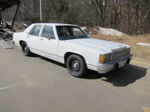 1991 Ford LTD Crown Victoria - This white sedan, likely from the early 1980s, is parked outdoors with a car carrier ramp nearby.
