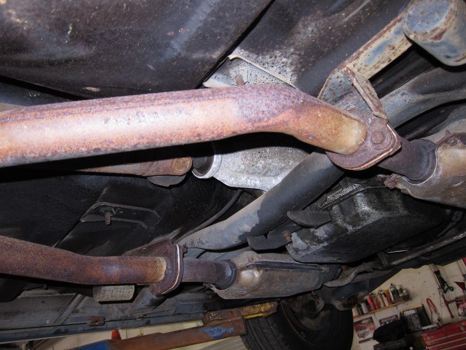 Underside view of vehicle showing exhaust system, transmission, and suspension parts. Some rust is visible on the exhaust