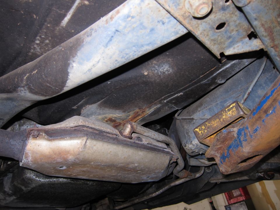 Underside view of a vehicle's exhaust system and frame components, showing rust and wear
