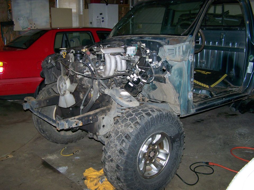 Truck body with engine bay open, front end disassembled. Red car in background
