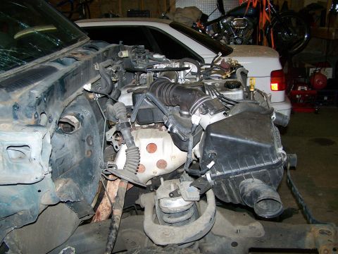 1988 Suzuki Samurai - The engine bay of a disassembled vehicle, showing the air intake system and engine block, with another vehicle and motorcycle in the backgr…
