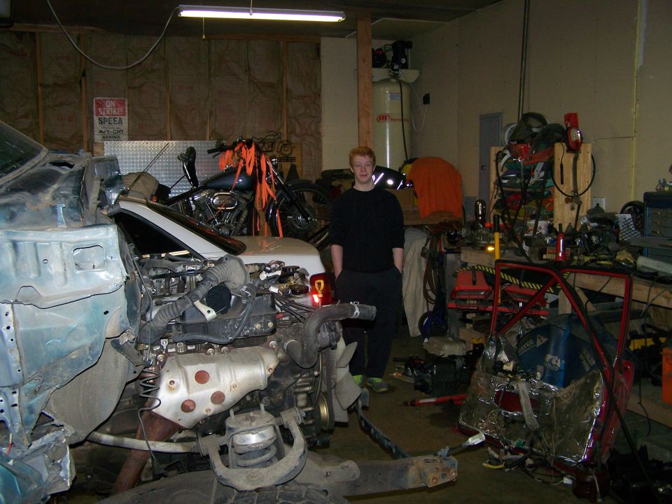 guy in garage with car engine and frame disassembled, motorcycle in background