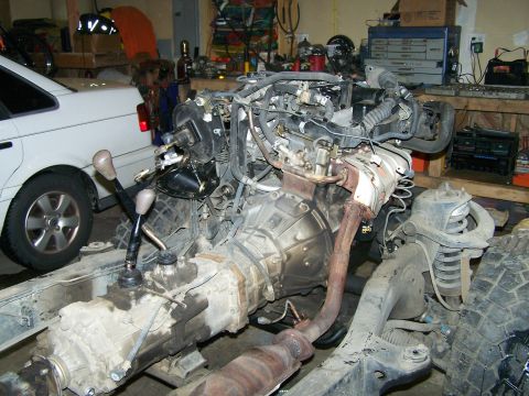 1988 Suzuki Samurai - Engine and transmission assembly with dual shifters, partially installed in a frame, with a white car in the background.