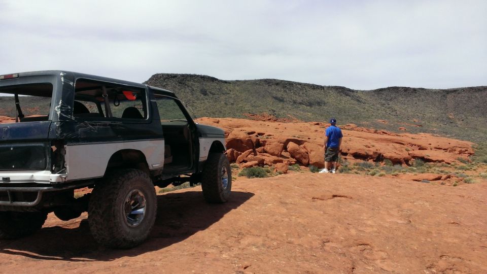 Modified Ford Bronco with large tires parked on red rock terrain, person stands in background