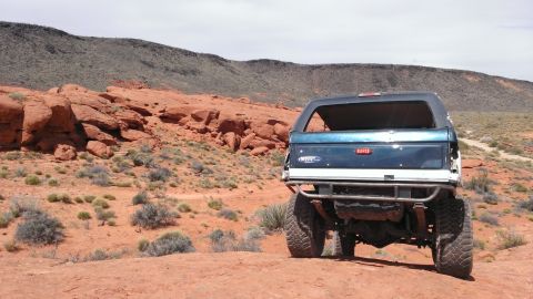 1994 Ford Bronco - A blue Ford Bronco is parked on a dirt road in a desert landscape with red rocks and sparse vegetation.