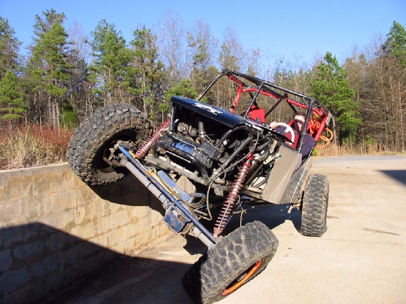 Tube-chassis rock buggy with red cage flexing front tire off a stone wall, winch and coilovers visible